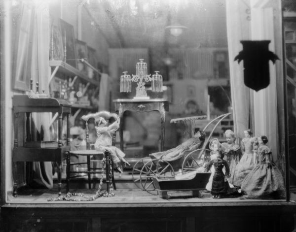 A nighttime view of dolls from the collection of Alice Kent Trimpey, arranged in the window of the Trimpey Studio and antique shop, 128 Fourth Avenue. The dolls are displayed with antique furniture and toys.