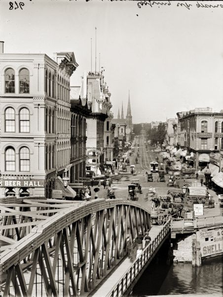 Elevated view over bridge and river looking up Grand Avenue. Pedestrians, horse-drawn vehicles and a street car are in the foreground.
