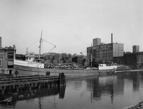 View across Milwaukee River of workers of the Herman M. Hettler Lumber Company unloading lumber off a shipping boat to the Shroeder Lumber Yards and Trostel tannery. Located on the west side of the Milwaukee River north of Pleasant Street bridge (in the left foreground).