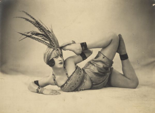 Billie Sidell contorts her body in a dancer's pose. She is wearing a beaded costume and a feathered headdress.
