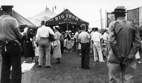 A crowd of people stand outside a circus tent. The words "Big Show" are emblazoned over the main entrance.