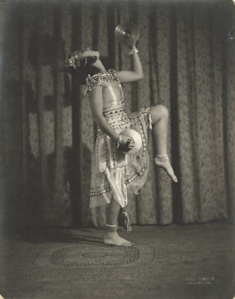 Pierre Sidell poses with cymbals, with her knee raised high. She is wearing an elaborate costume.