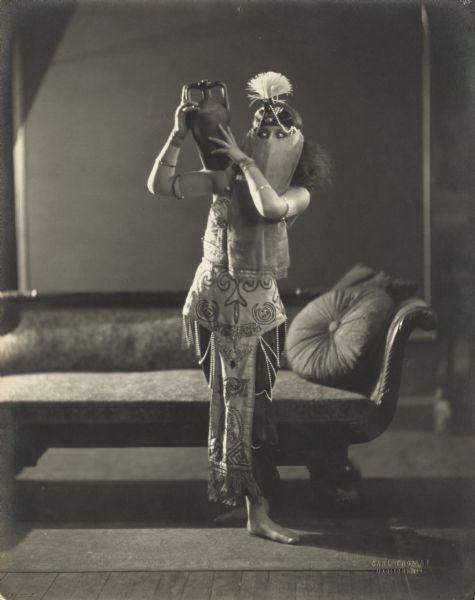 Pierre Sidell poses holding a small jug on her shoulder. She is wearing a face veil and headdress.

From an undated (but probably 1922) article in the Kehl School of Dance scrapbook (M92-249):
"Burning sands of the desert; spicy fragrance of Araby; persimmon moons that shine on gardens of the East; the dance of a slave girl for her master--all will be woven into the dance prologue to be given with the return showing of the Sheik at the Majestic [movie theater in Madison, Wisconsin].
"Miss Pierre and Billie Sidell, Central high school students, will give the dance of the Sheik, Pierre as the slave girl and Billie as the Master.  They are students of Leo Kehl.
"The Sheik costume is modeled from that worn by Rudolph Valentino of the picture.  The silken surfaces of the slave girl's harem dress will catch the rosy lights flickering over the dancers as they portray the desert romance."
