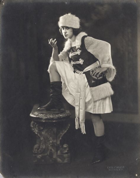 Pierre Sidell wears a white fur hat and tunic with a fur collar. She is holding a cigarette as she poses with one foot up on a bench.
