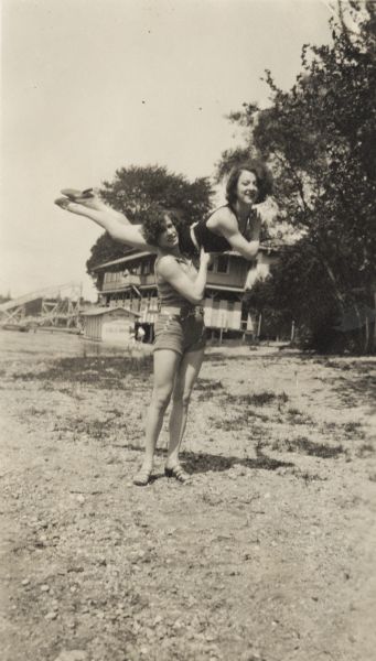Billie (Violet) Sidell holds her sister Pierre Sidell aloft over her shoulder in a pose. They are posing outdoors on a shoreline wearing what appears to be bathing suits. In the background is a two-story building and a large structure that might be part of an amusement park.