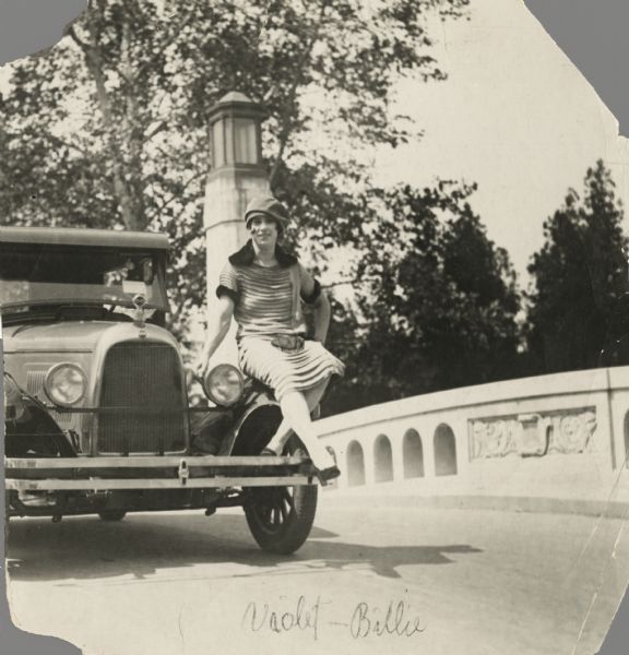 A fashionably-attired Billie (Violet) Sidell poses seated on the front fender of an automobile, which appears to be parked on a bridge.