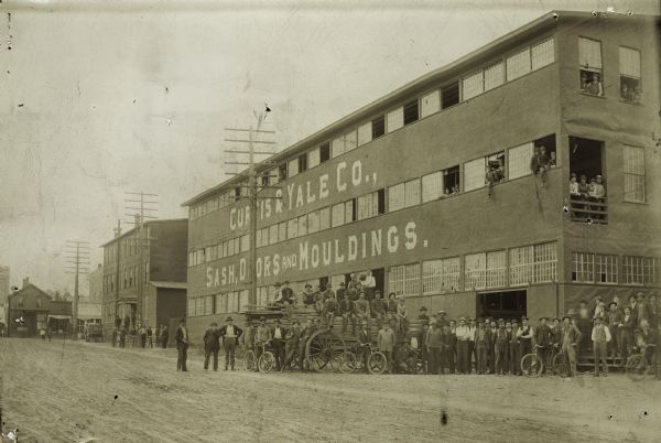 Group of men posing in front of Curtis and Yale Co. Plant one. A number of workers are looking out from windows in the building. Other workers pose on a wagon loaded with wood, and some stand with bicycles.