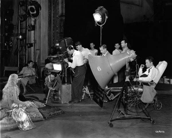 Production still during the filming of "Citizen Kane." Orson Wells smokes a pipe and directs Dorothy Comingore (playing Susan Alexander Kane) from a wheelchair. Cinematographer Gregg Toland, wearing a visor and sunglasses, stands directly behind the blimped camera along with other technicians.