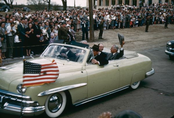 President Harry S. Truman in Madison for the opening and dedication of the Filene House, 1617 Sherman Avenue. He is sitting in the car and waving his hat during a parade. Spectators are standing along the side of the road. Filene House was the headquarters of the Credit Union International Association (CUNA).