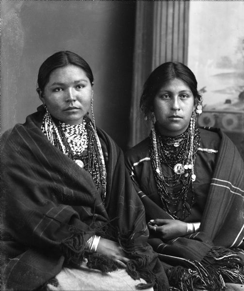 Waist-up studio portrait of two Ho-Chunk women posing sitting in front of a painted backdrop. They are wearing several necklaces, earrings, file bracelets, and shawls over their shoulders. Both women are wearing a photographic pendant on their necklaces. They are identified as Caroline Decorah Black Deer on the left, and Mabel White St. Cyr on the right, who is wearing Winnebago chain and coin earrings.