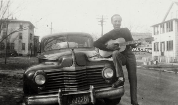 Lewis Arms strums a guitar, with one foot resting on the bumper of a car at 629 Milton Street.