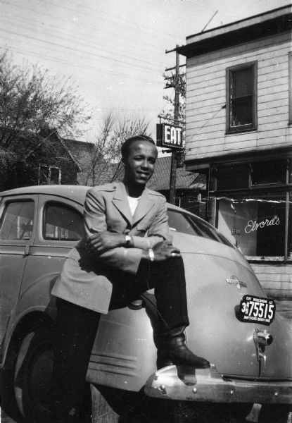 Lewis Arms poses next to an automobile in front of Carl Elvord's Barbeque restaurant on the corner of Mound and Murray Streets in "the Bush".