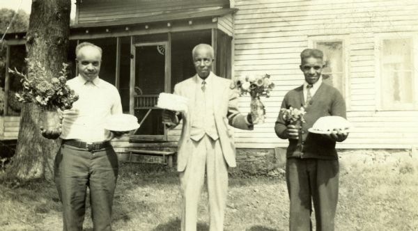 Three men from the Shivers family pose holding cakes and vases of flowers. One of the men is Tom Shivers and one is Algie Shivers, and the other man is unidentified. They are standing outside and a house with a porch is in the background.