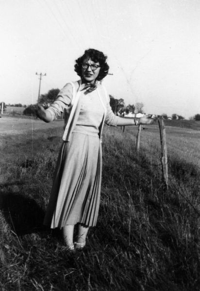 Carol Commack Banks stands at a roadside in the grass next to a post fence. There are houses in the far background.