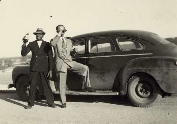 Joe Brown and Lewis Arms pose next to an automobile wearing dapper suits and hats. They are drinking from bottles.