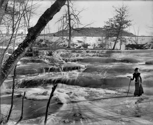 A well-dressed woman with a walking stick stands on ice at the partially frozen Skillet Falls. Bare trees frame the scene. There are patches of snow and tree stumps on a hill in the background.  The hill is known locally as Mount Baldy.