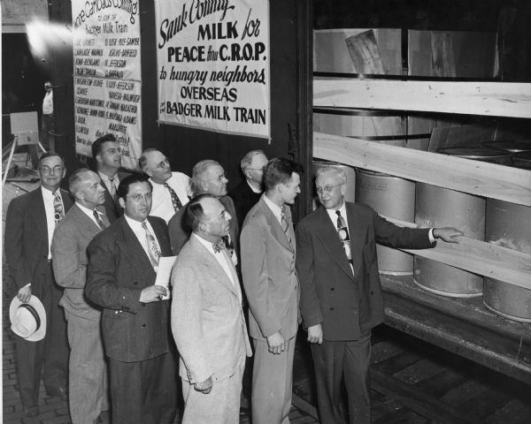 A group of men are standing next to the Badger Milk Train, which was part of the Sauk County Milk for Peace program. As the sign behind them notes, this program got milk "to hungry neighbors overseas". The men in the front row are, from left to right: J.B.F. Hayes, Erich Lenz, Noble Clark, Bob Lewis, and Governor Rennebohm. In the back row, from left to right, are Claire L. Jackson, Warren Clark, Reverend Sorenson, and Father Hausler.