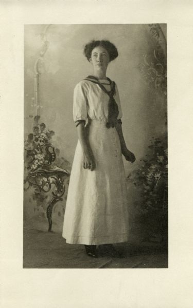 Photographic postcard of a full-length studio portrait of Miriam Bennett in front of a painted backdrop with postcard conventions on back.