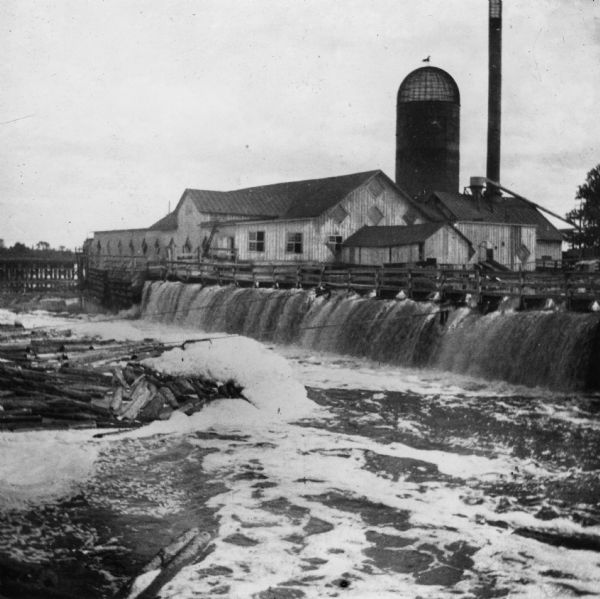 View of the Peshtigo Company sawmill including the old wooden dam in the foreground. There are logs in the water on the left.