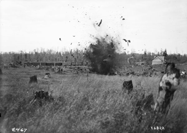 View of a stump as it's being blown up with explosives in a field on a farm. In the background is a train.
