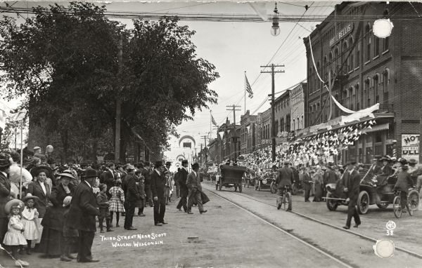 View looking South down Third Street near Scott. The street is filled with a large crowd, bikes and automobiles. Banners and flags are along the sidewalk in front of storefronts. Caption reads: "Third Street Near Scott, Wausau Wisconsin".