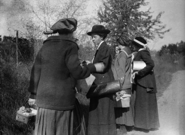 Four women from Rockford College standing by the side of a dirt road looking into a wooded area. The women are carrying picnic baskets.