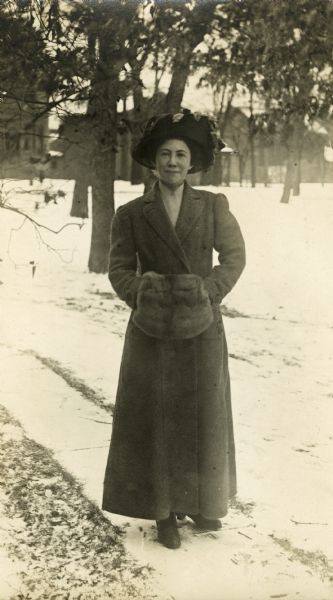 Photographic postcard of a full-length portrait of Miriam Bennett outdoors in front of the Bennett house on a winter day. A light layer of snow is on the ground and Miriam is wearing a long coat, a decorated hat and a fur muff.
