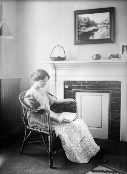 Unidentified woman seated in a wicker chair reading in the Jane Adams room at Rockford College. A walled-over fireplace is on the wall behind her, and an H.H. Bennett photograph of the Wisconsin Dells hangs above the mantle.