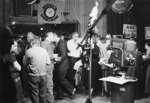 People mill about the WKOW television studio. A television camera and boom microphone are visible to the right, and there is a clock in the background surrounded by the phrase "Today's Farm & Home".