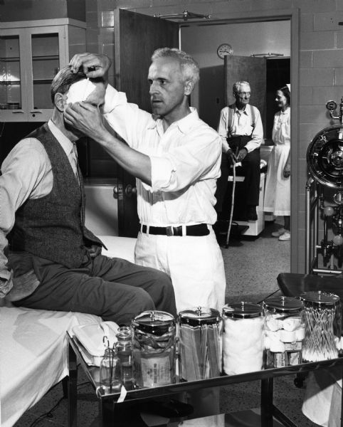 A doctor bandages a man's head at the Dane County Hospital and Home. Another patient waits in the background with a nurse.