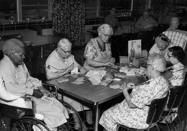 A group of elderly women sit together at a table working on March of Dimes activities. Pictured are Ona Smith, Julie Simonson, Mary Zweifel, Gussie Rowe, Minerva Holland, and Stella Anderson. Men and women sit in the background at tables.