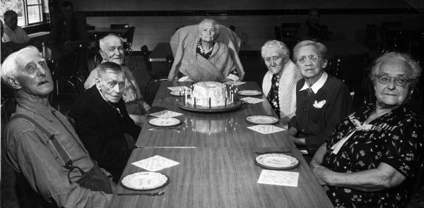 Seven residents of the Dane County Hospital and Home gather around a table to celebrate a birthday. A birthday cake sits on the table.