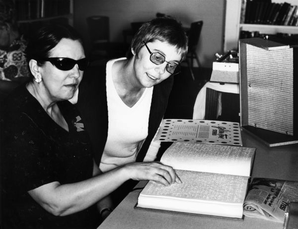 A woman in dark glasses, who is a resident of Dane County Hospital and Home, reads a book in Braille as her therapist looks on.