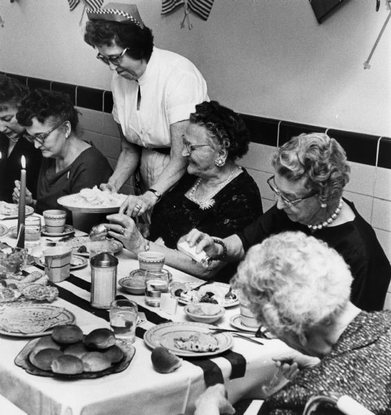 Several women are served lutefisk at a lutefisk supper at the Dane County Home. There are plates of lefse and dinner rolls on the table.