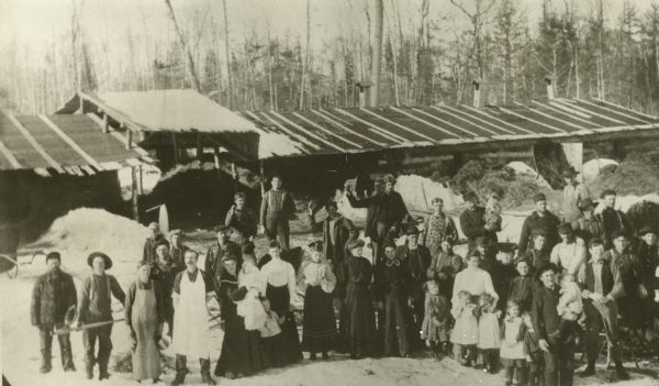 Elevated view of lumberjacks joined by their wives and children on a Sunday at the Auburndale Lumber Camp.