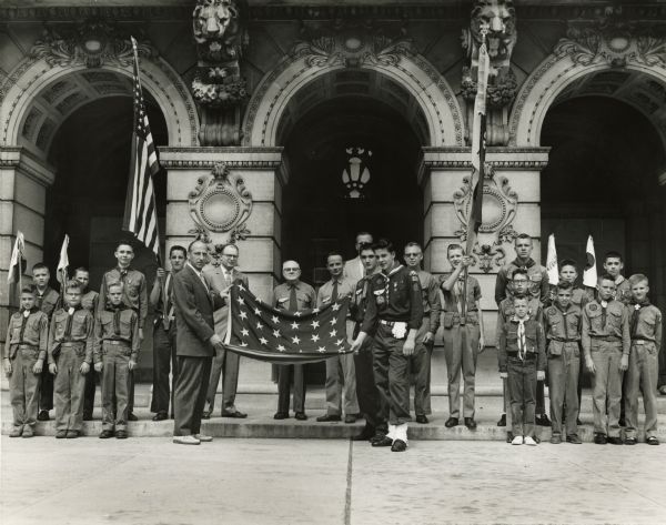 Leslie H. Fishel, Jr. participating in a ceremony in front of the State Historical Society of Wisconsin with a large group of Boy Scouts and Boy Scout leaders. He and a Boy Scout are holding a flag. He was the Director of the State Historical Society of Wisconsin from 1959 until 1969.