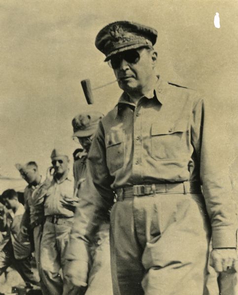 General MacArthur in uniform with corncob pipe. Unidentified military personnel are visible in the background.