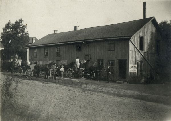 Men with horses and wagons gathered outside the Faville Grove Farmers' Co-Operative Cheese Factory. A poster advertising the State Fair is pasted to the wall of the building.