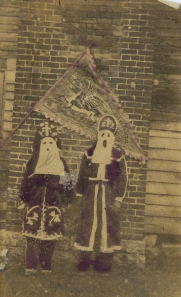 Two people in robes and hoods standing beneath a triangular flag with an image of a dragon on it. The robes and part of the flag are hand-colored in purple.