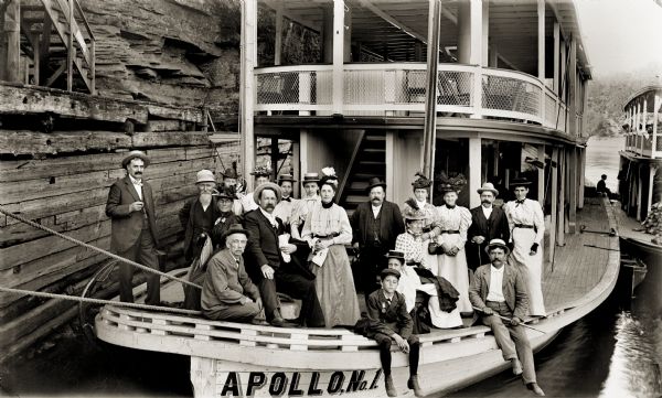 View of bow of "Apollo No. 1" steamboat with passengers posing near a dock on the shoreline.