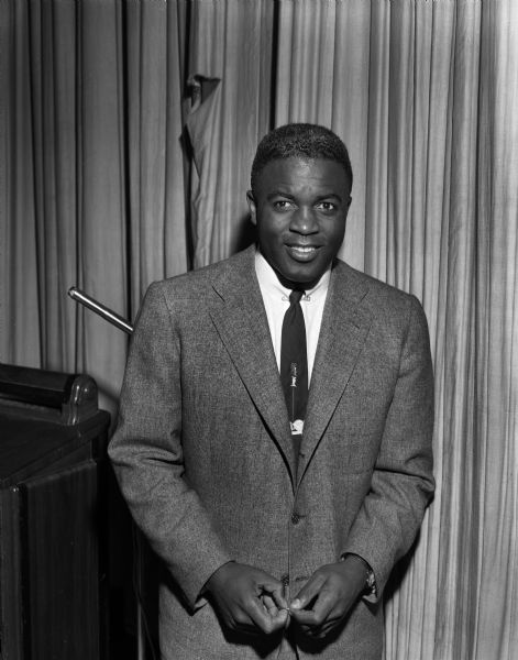 Jackie Robinson standing in front of a curtain wearing a jacket and tie. He is standing next to a podium, perhaps behind the curtain before or after speaking.