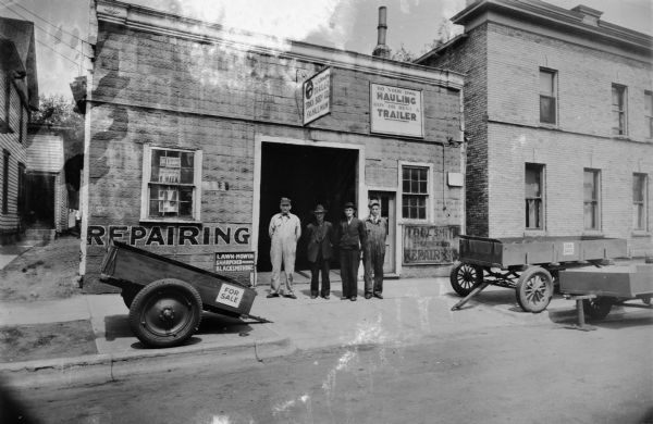 View of exterior of Trailer Truck and Body Co. repair services. Four men are posed in front of garage. One man is wearing a suit, the others wear work clothes. Sign above is Madison Trailer Truck Body Co. F.A. Hallman. Other signs: "Repairing," "Do Your Own Hauling, Buy or Rent a Trailer," "Toolsmith and General Repairing," "Lawn-mowers Sharpened-Repairs, Blacksmithing" and "For Sale."