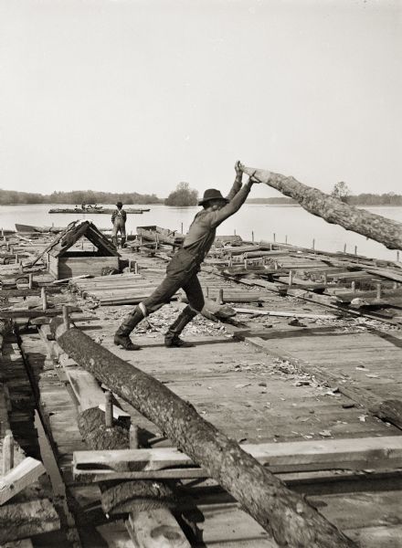 A raftsman at work raising an oar (possibly rowing) with another man standing in the background. A second raft is in the distance.