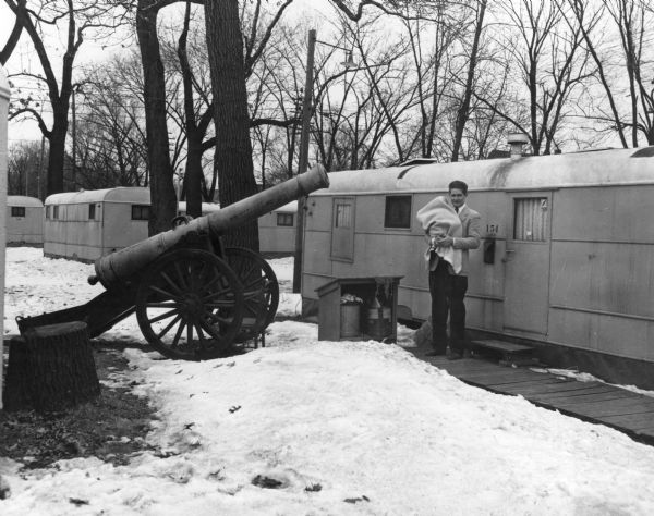 A University of Wisconsin veteran holding his baby outside of his Camp Randall trailer camp house. There is a large cannon pointing at him and his trailer.