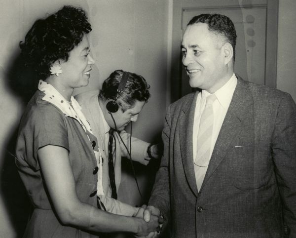 Daisy Bates shakes hands with Ralph J. Bunch, Secretary General of United Nations. A man wearing a headset is behind them.
