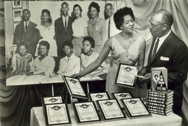 Daisy Bates speaking with unidentified man wearing eyeglasses. Copies of her memoir "The Long Shadow of Little Rock", and plaques from the NAACP for the nine students who integrated Central High School in Little Rock, are on a table. A large picture of Daisy and the nine students hangs in the background. They are, left to right, front row: Thelma Mothershed, Minnijean Brown, Elizabeth Eckford, Gloria Ray; and row 2, left to right: Jefferson Thomas, Melba Pattillo, Terrence Roberts, Carlotta Walls, Daisy Bates, and Ernest Green.