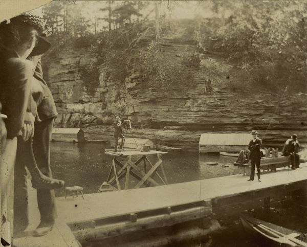 Henry Hamilton Bennett stands with his camera on a platform. He built the platform in order to take photographs of passengers in the boats that toured the Wisconsin River.