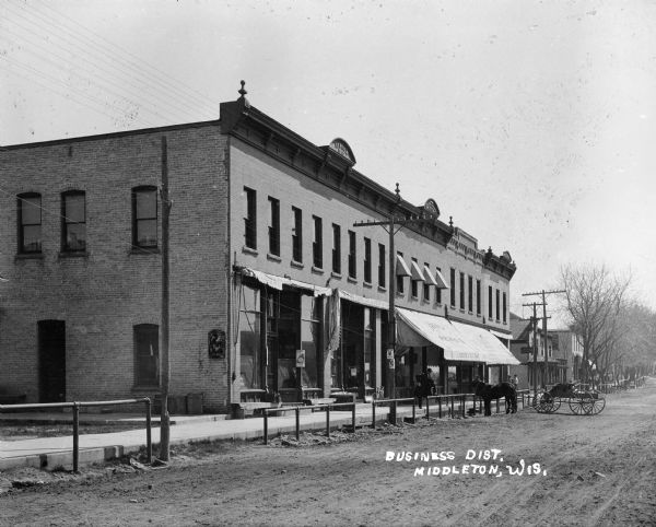 View from road of the business district, with a horse-drawn wagon parked in front of a large commercial building near the sidewalk. The first of three decorate arches on the facade of the building at the roof has carving that says: "Heprien 1900".