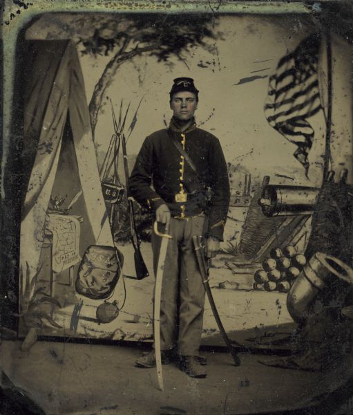 A tinted tintype of a Civil War soldier posing with an officer's sword in front of an elaborately painted studio backdrop depicting a tent, an American flag, and a cannon.  The soldier is possibly Stanley Lathrop from Montello who served in the 1st Wisconsin Cavalry.