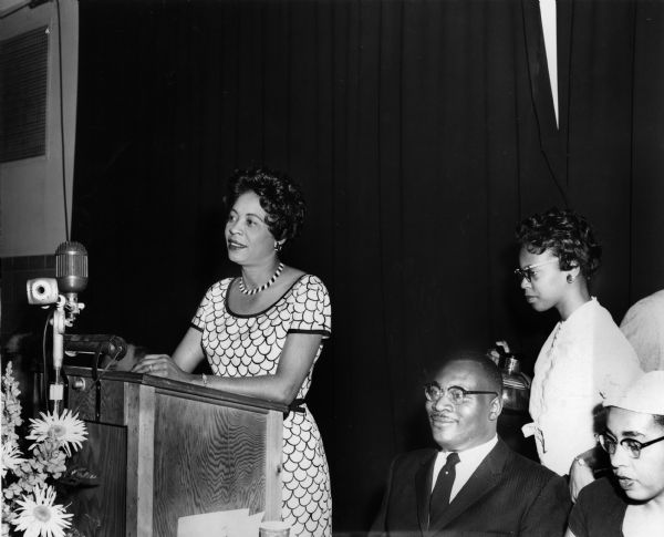 Daisy Bates speaks at a podium, with several people, including a smiling man, seated to her left.